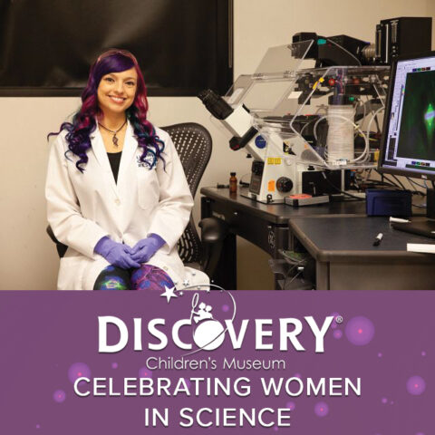 woman in a lab coat with colorful hair sitting at a desk with scientific equipment