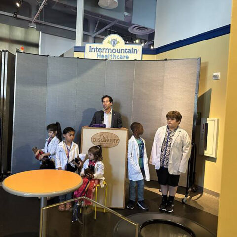 Hombre dando un discurso en un podio rodeado de niños con batas de laboratorio en el Discovery Children&#039;s Museum.