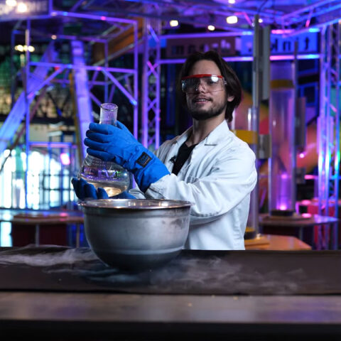 un hombre barbudo con bata de laboratorio que sostiene un gran vaso de precipitados lleno de líquido junto a un gran cuenco metálico en el Discovery Children&#039;s Museum