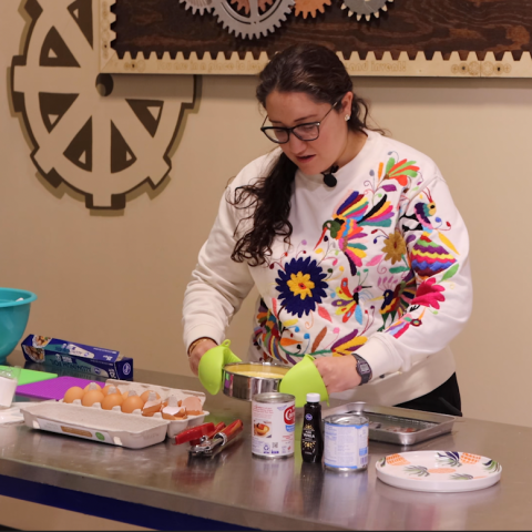 A cooking instructor holds a circular metal pan full of batter over a table scattered with flan ingredients and cooking tools