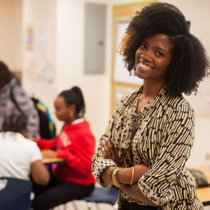 a woman smiles at the camera while standing near students at their desks