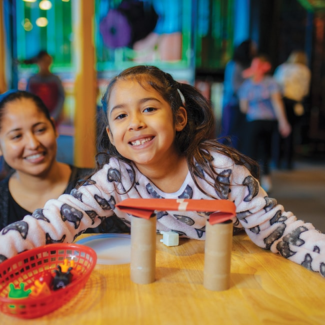 A child smiling at the camera with her mother looking at her in the background