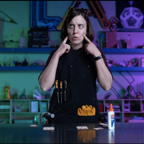 a woman wearing an apron full of tools looks up quizzically while touching her pointer fingers to her cheeks