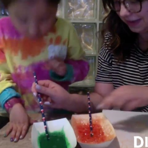 an instructor pointing to a dish with bubbly paint while holding a straw in another dish of paint in front of a child