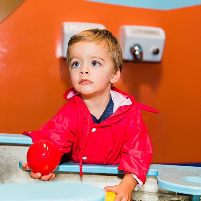 赤いレインコートを着た幼児が赤と黄色の小さなボールを手に持っている。