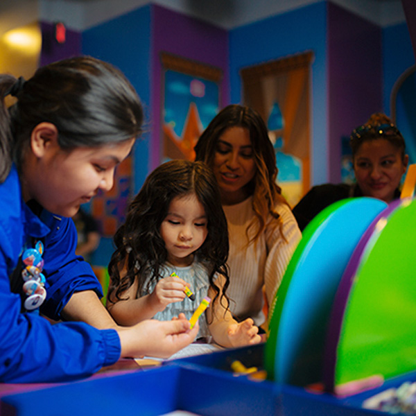 Una joven educadora del museo ayuda a una niña con lápices de colores mientras su madre y una amiga miran al fondo.