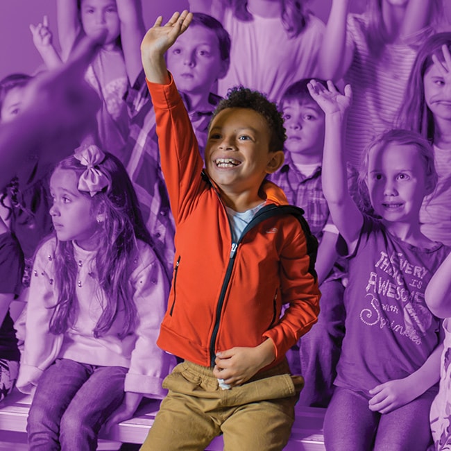 In full color and focused, a young boy with a raised hand, enthusiastically waiting to be called and around him are his young peers, some also have their hands raised