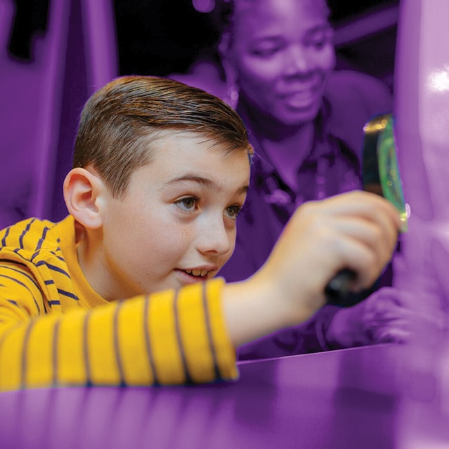 in full color and focused, a young boy is holding a magnifying glass whilst his teacher looks on