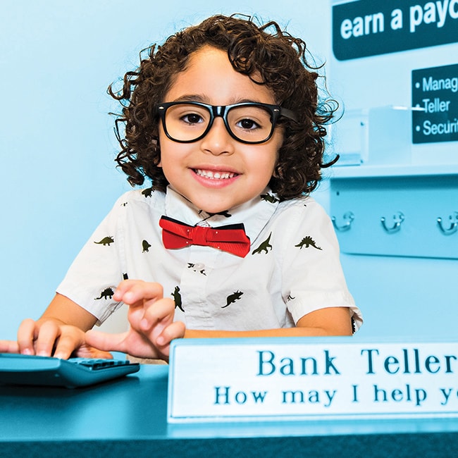 young boy wearing glasses, a red bowtie, and dinosaur collared shirt smiling at the camera and pretending to be a bank teller