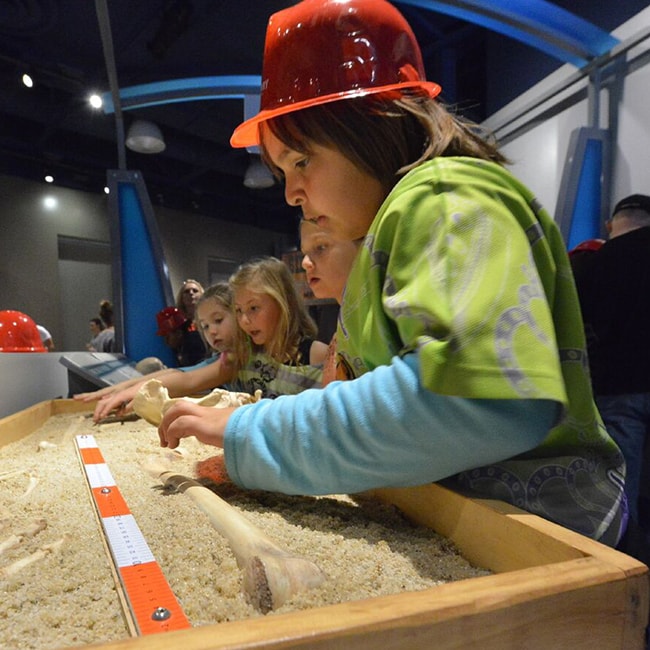 A child in the foreground wearing a plastic hardhat helmet standing over a mini construction site that has sand