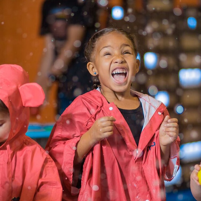 A young girl wearing a red raincoat and laughing as she gets splattered wet inside the exhibit Water World