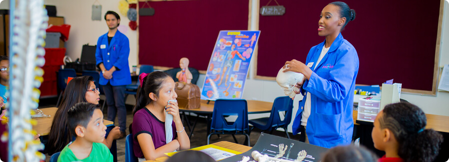 Un docente adulto con la bata azul del Discovery Kids Museum haciendo una presentación delante de los niños en un aula.
