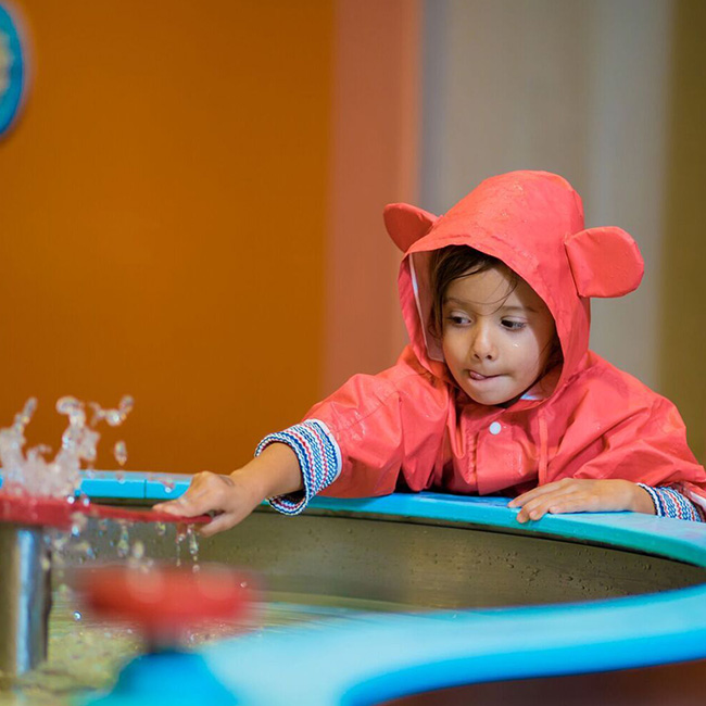 A toddler reaching over to a water feature inside the museum while wearing a red raincoat with bear ears