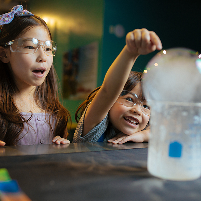 Dos niñas con gafas de seguridad admiran una bola de hielo circular sobre un vaso de precipitados.