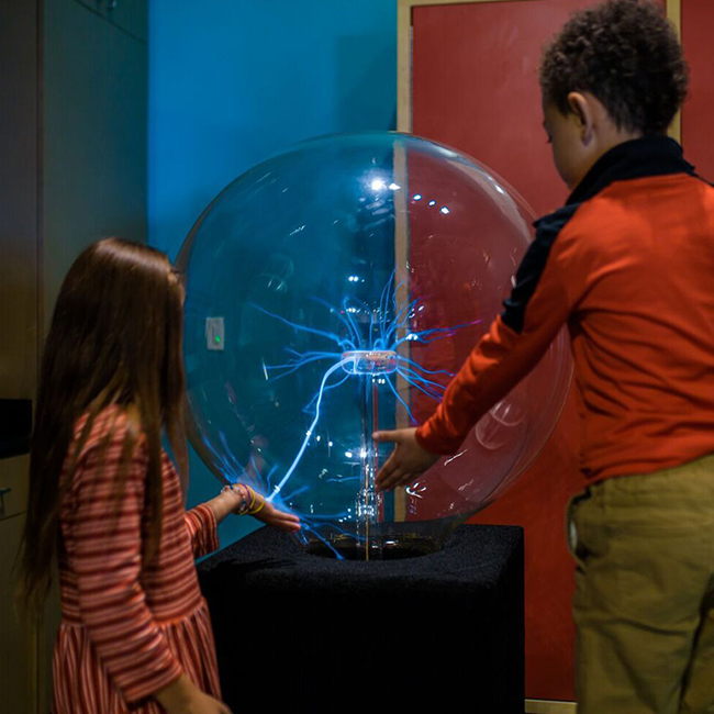 a young girl and young but taller boy holding onto and standing in front of a large plasma ball