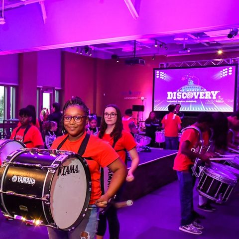 young woman with drum strapped to her body in a purple lit room with other members of a youth drum line at the Discovery Children's Museum