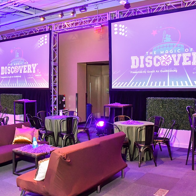 un espacio abierto para eventos con mesas, sillas, sofás y 2 pantallas de proyección en las que se puede leer &quot;La magia del descubrimiento&quot; en el Discovery Children&#039;s Museum