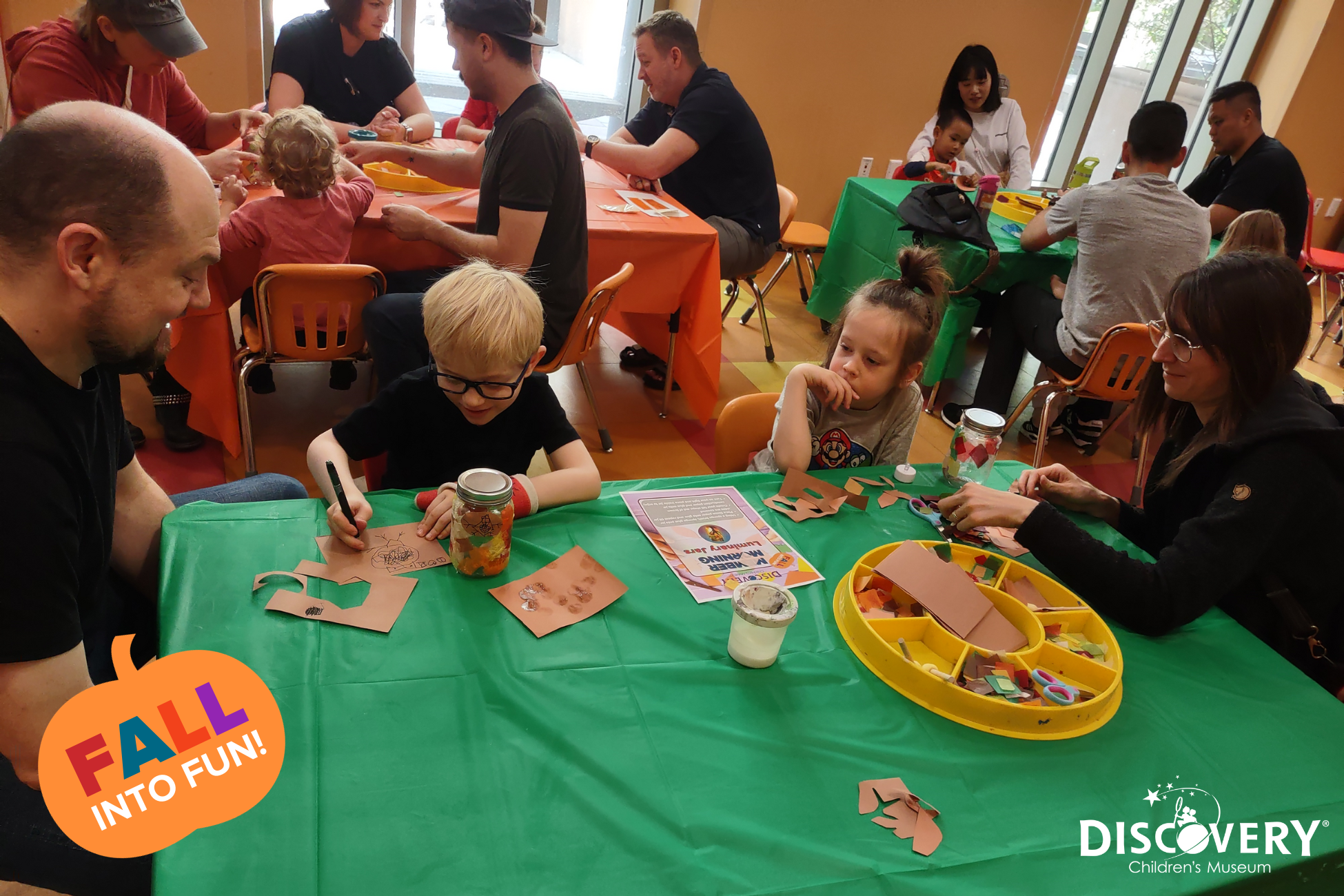 2 padres y un niño observan a un niño dibujar en una cartulina en una mesa de manualidades verdes del Discovery Children&#039;s Museum.