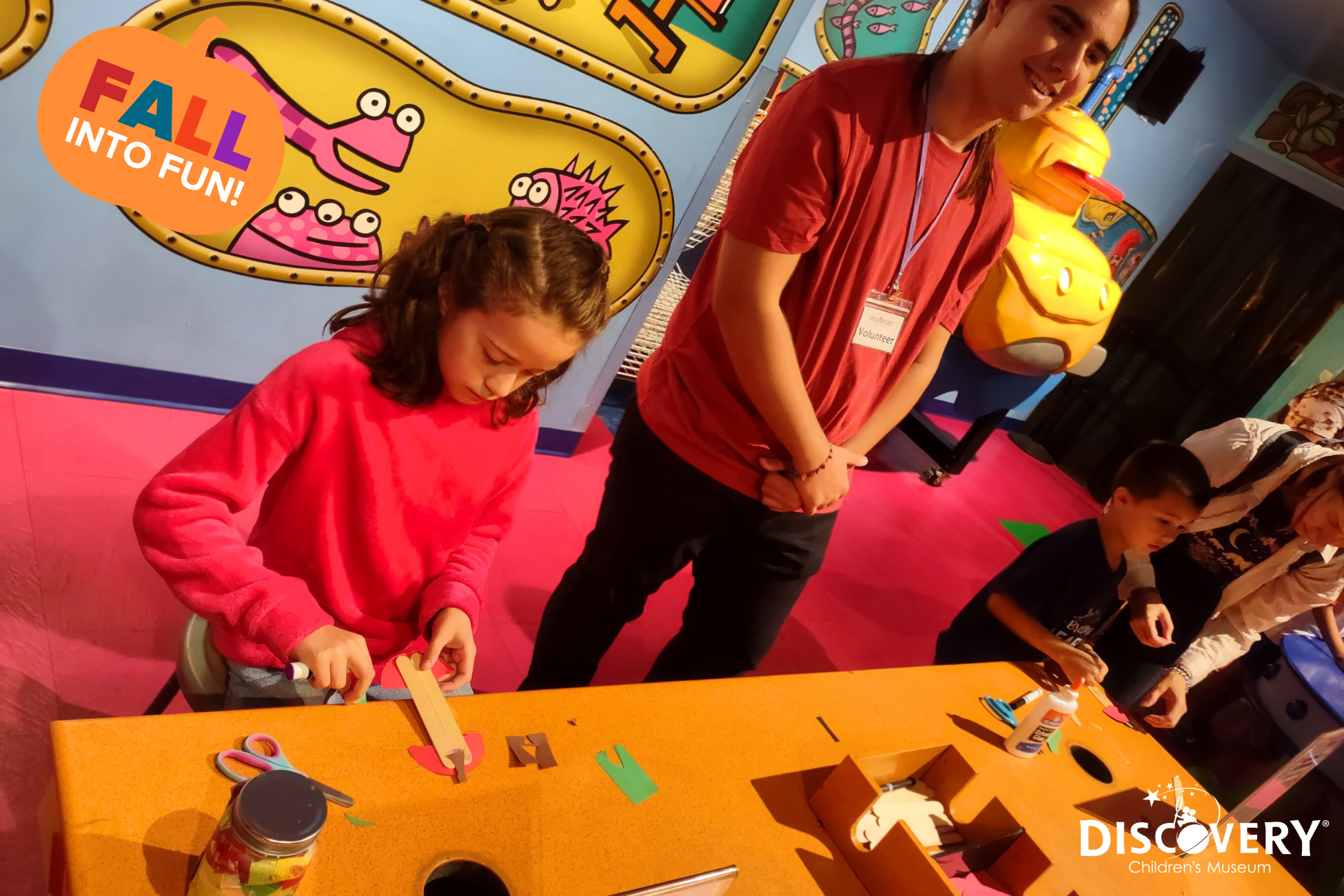 在发现儿童博物馆，一名博物馆志愿者站在一名用纸和胶棒制作手工艺品的小女孩身旁。