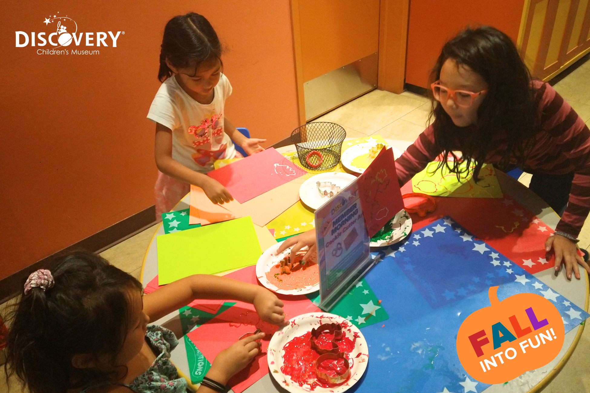 3 名儿童在发现儿童博物馆一张色彩斑斓的桌子前，将饼干切割器浸入颜料中