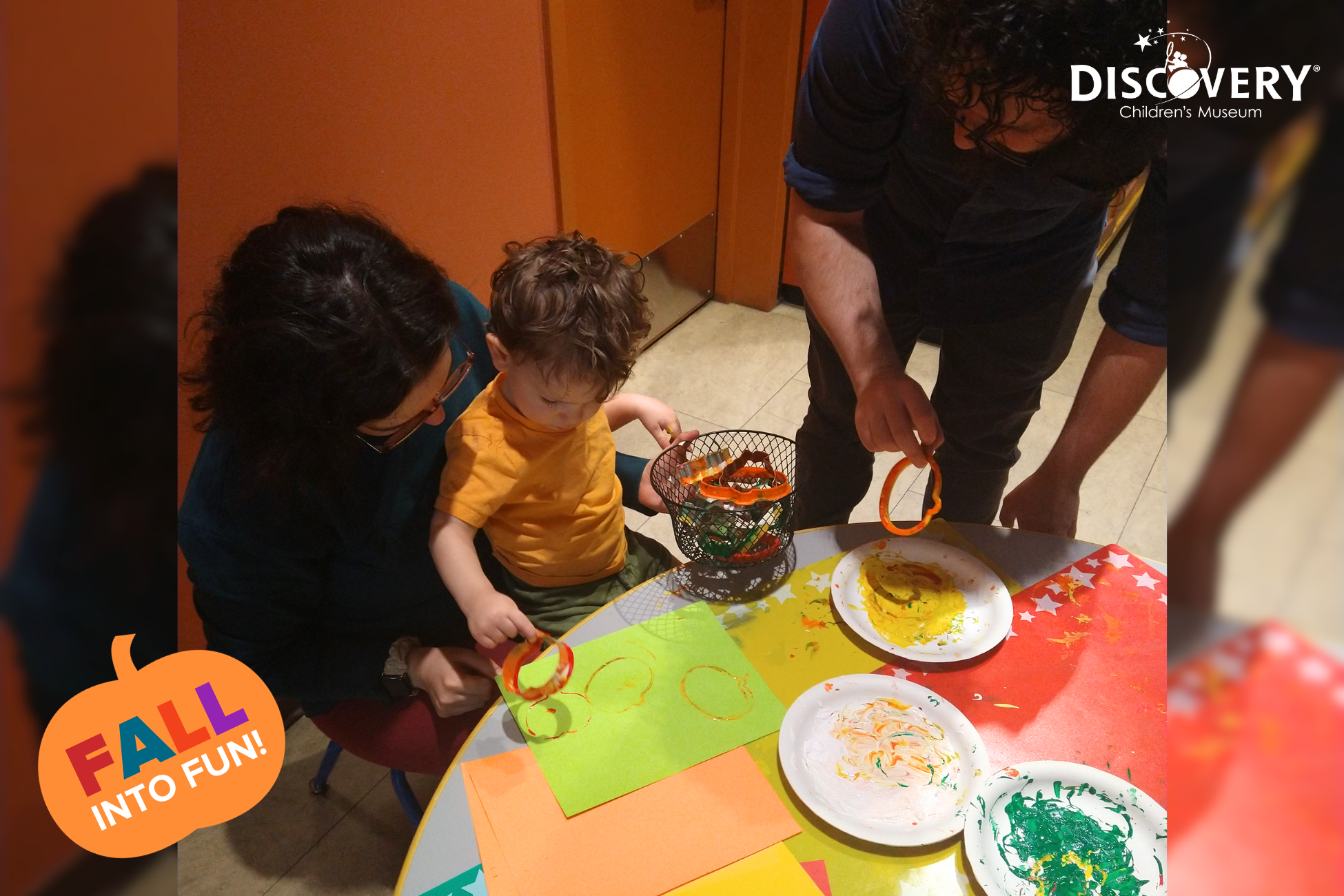 在发现儿童博物馆，一个孩子坐在母亲腿上，和父亲一起用饼干刀和颜料制作图案