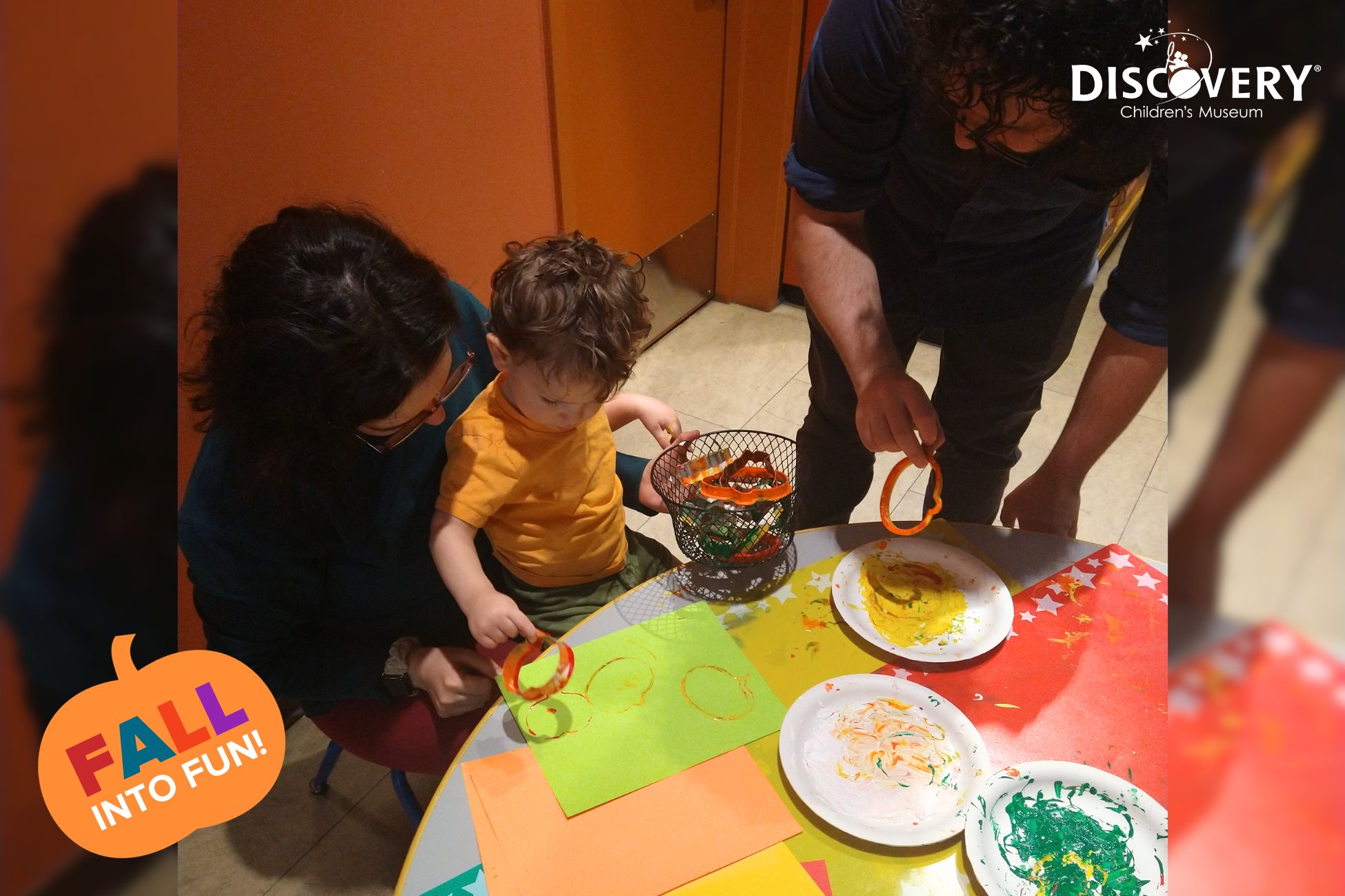 un niño sentado en el regazo de su madre usando cortapastas y pintura para hacer diseños con su padre en el Discovery Children&#039;s Museum