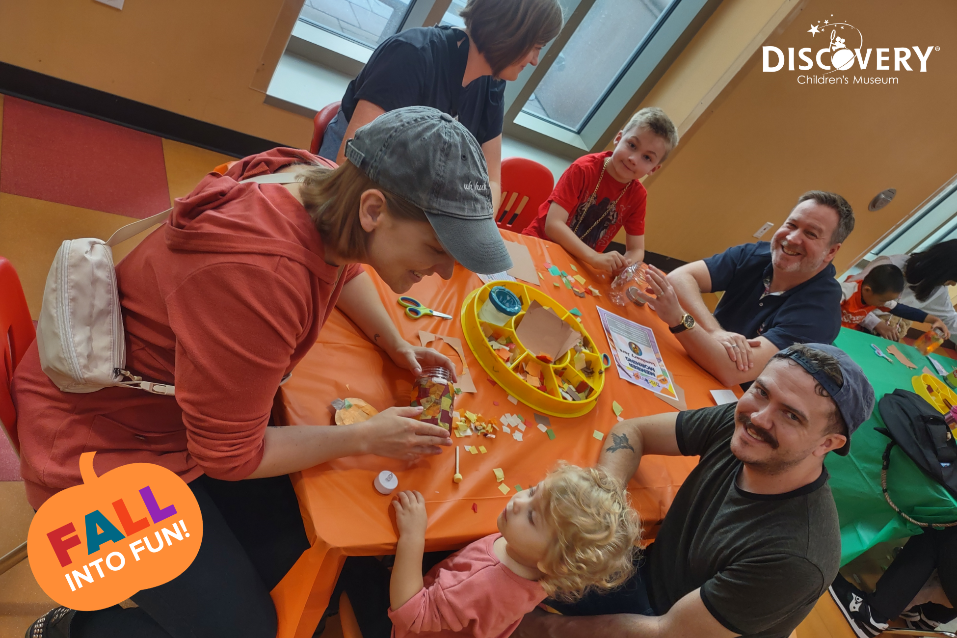 4 名家长和 2 名儿童坐在发现儿童博物馆的橙色小桌旁，桌上摆放着一些彩纸