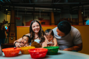 ディスカバリー・チルドレンズ・ミュージアムのテーブルで、幼児のお絵かきを見ている男性の隣で、笑いながらポーズをとる女性と子ども。
