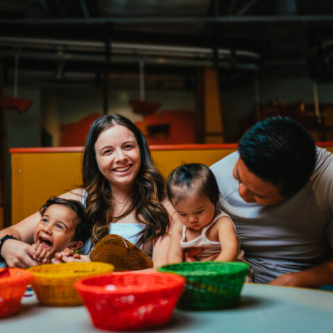 una mujer y un niño ríen y posan sentados junto a un hombre que observa a su hijo pequeño dibujar en una mesa del Discovery Children&#039;s Museum