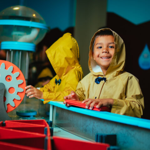 child in a raincoat smiling at the camera and standing by an interactive exhibit at the Discovery Children&#039;s Museum