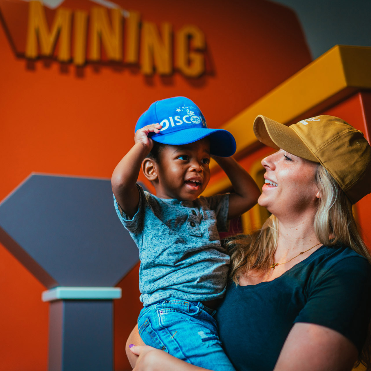 a woman holds up a toddler boy who is putting on a ball cap at the Discovery Children's Museum