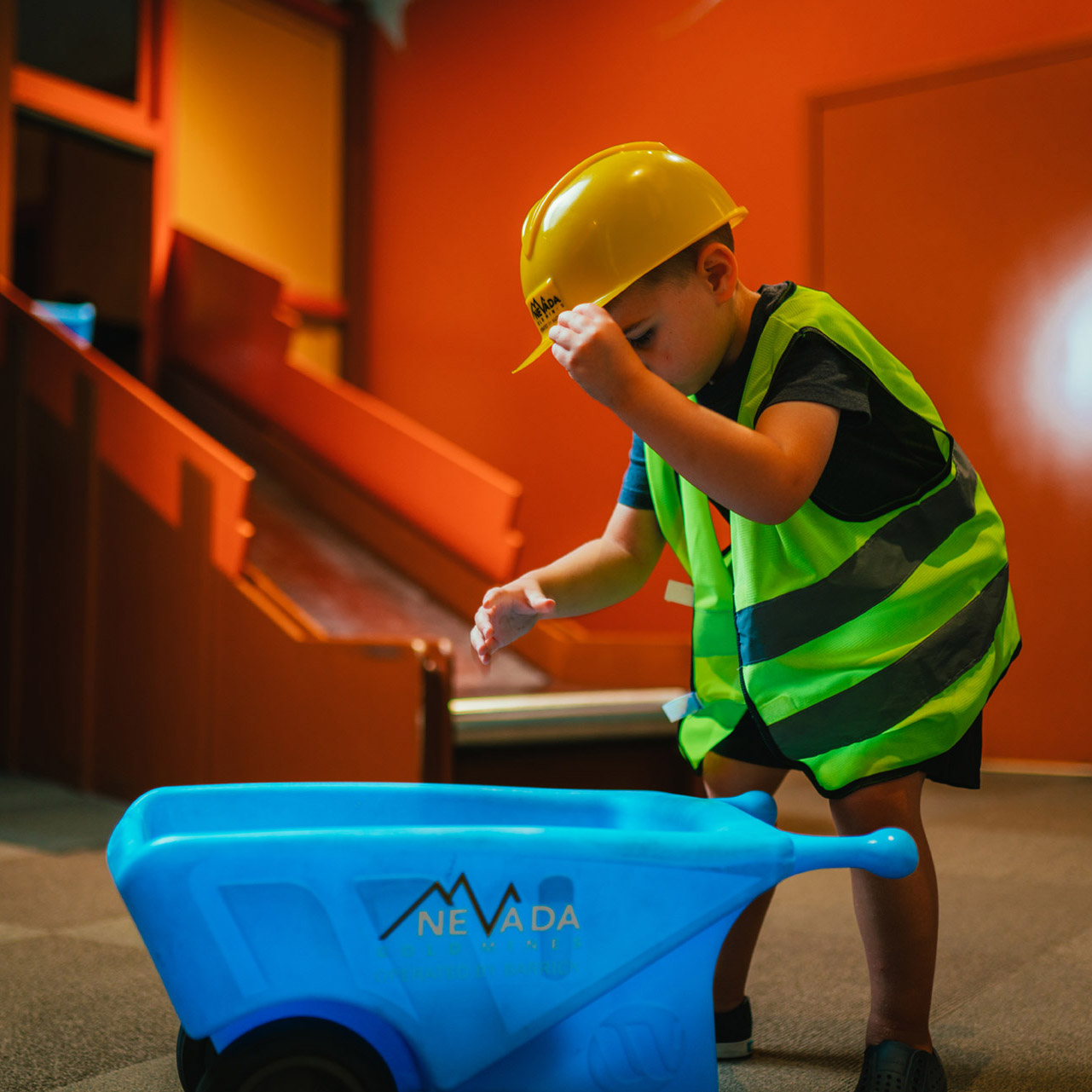a toddler in a hart hat and reflective vest stands over a small plastic wheelbarrow at the Discovery Children's Museum
