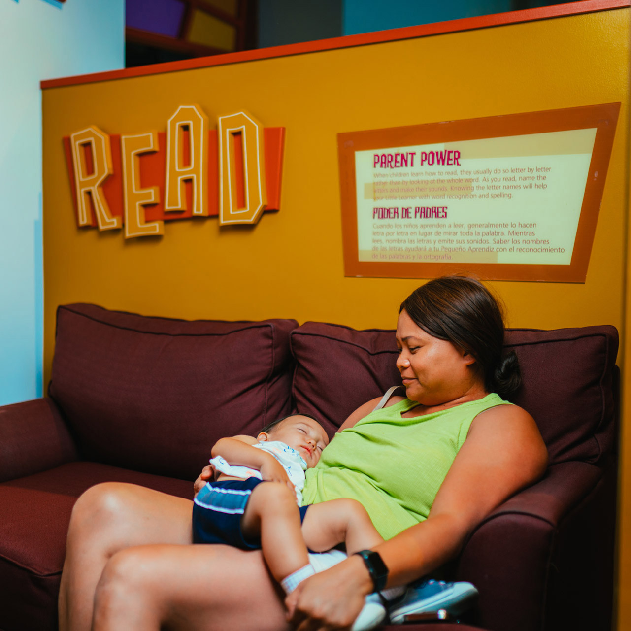 ディスカバリー・チルドレンズ・ミュージアムの &quot;read &quot;と書かれた看板の前で、ソファに座った女性が膝の上に寝ている幼児を抱いている。