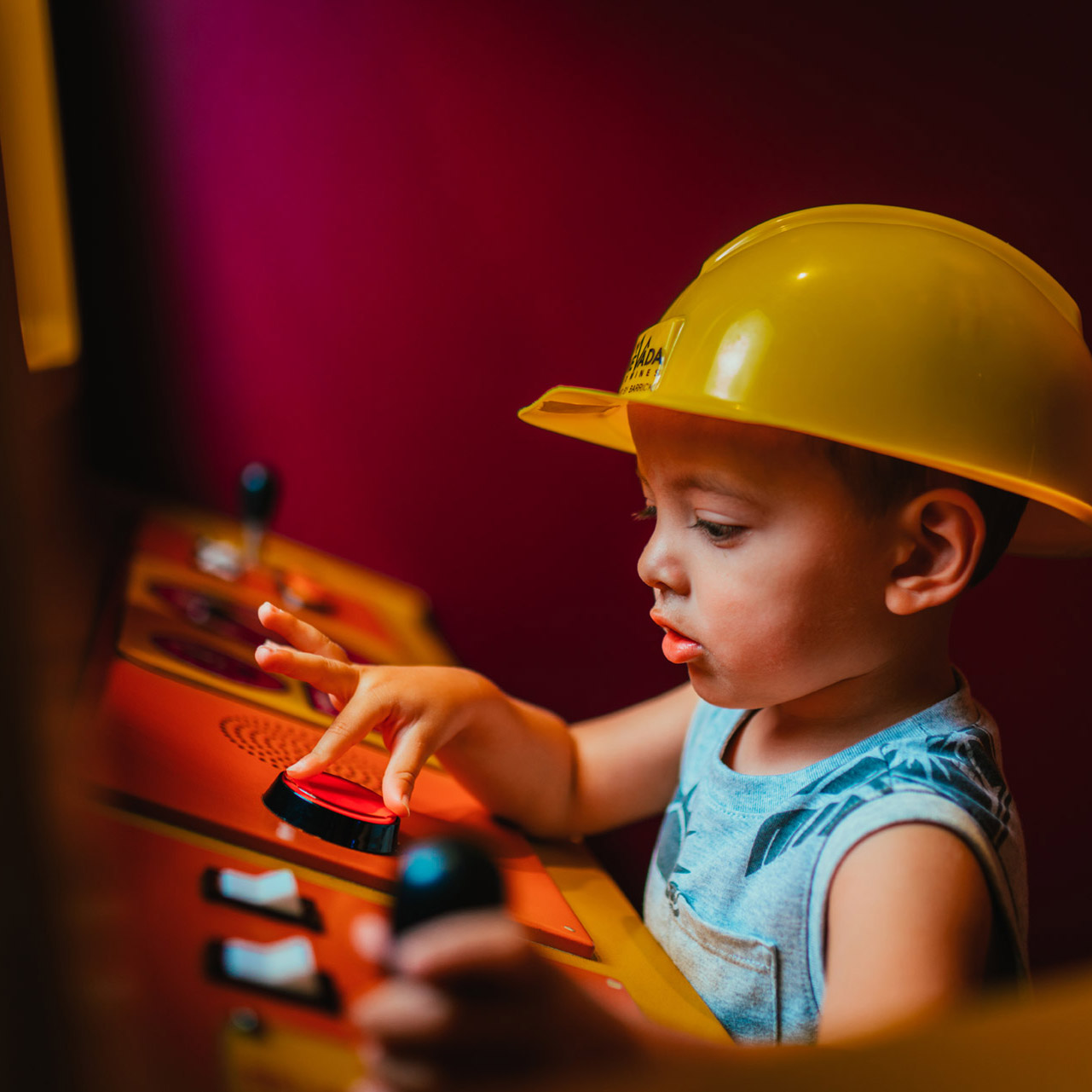 一个戴着坚硬帽子的学步男孩按下发现儿童博物馆控制台上的红色按钮